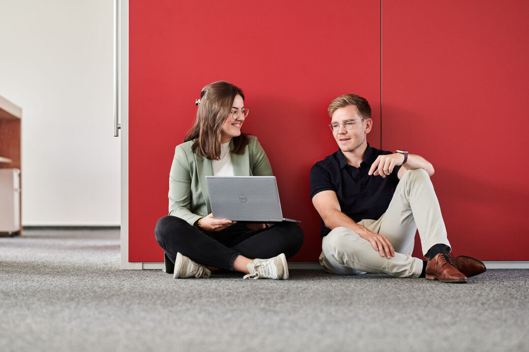 d.velop Mitarbeitende mit Laptop auf dem Fußboden vor roter Wand